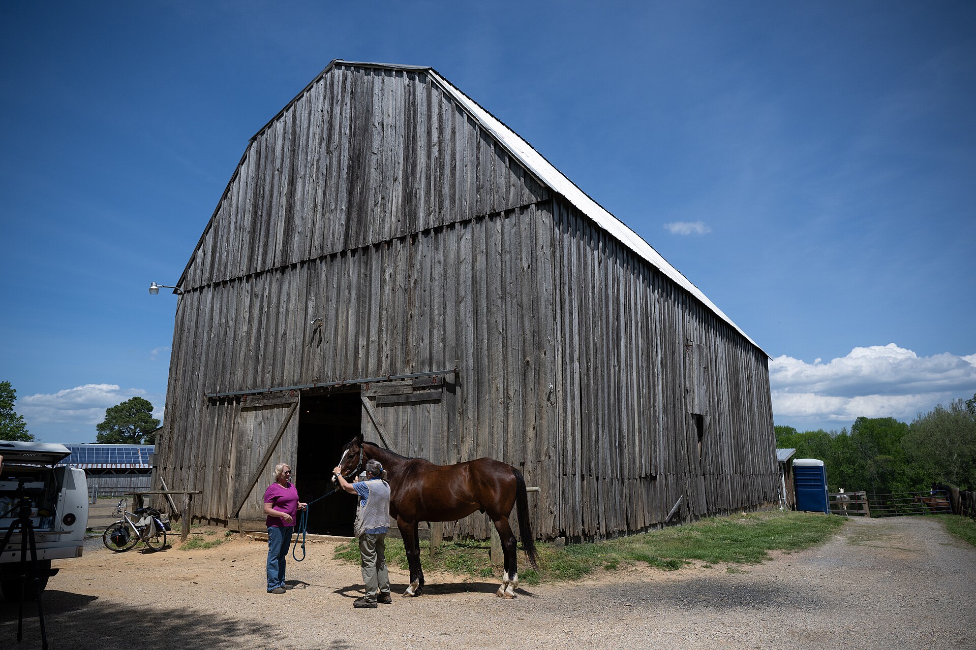 A veterinarian and a bay horse stand outside a large wooden barn.